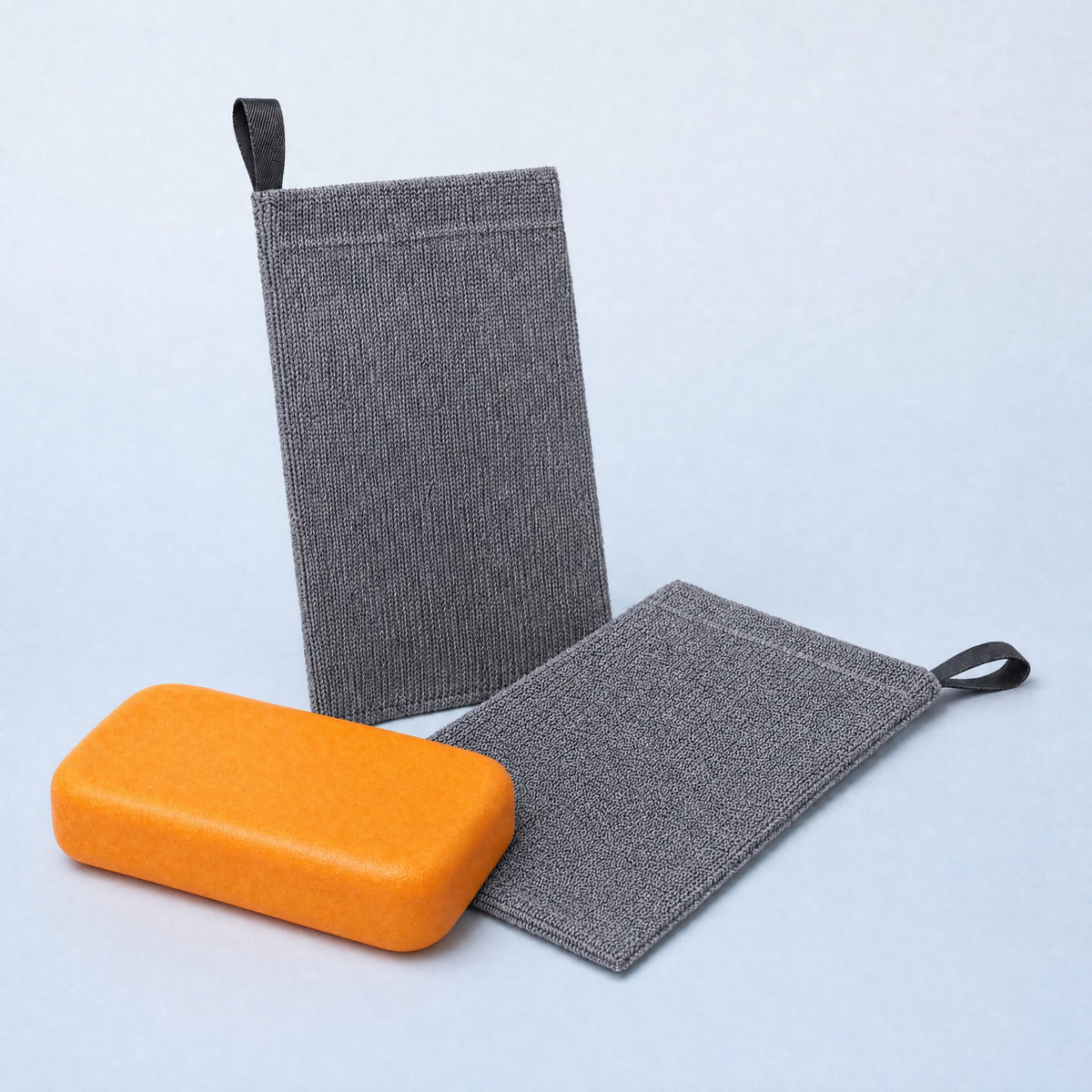 Two grey textured Wakai exfoliating soap bags with black hanging loops beside an orange rectangular Wakai bar