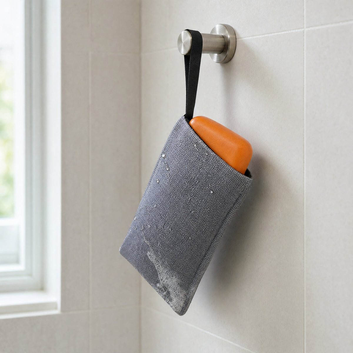 Grey exfoliating soap bag holding an orange Wakai bar, hanging from a shower hook